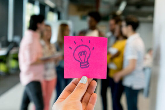 Hand holding up a bright sticky note with a lightbulb drawn on it in an innovative office space.