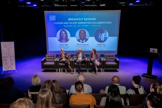 Three people speaking at a breakout panel at a conference with dim lights and a big screen behind them.