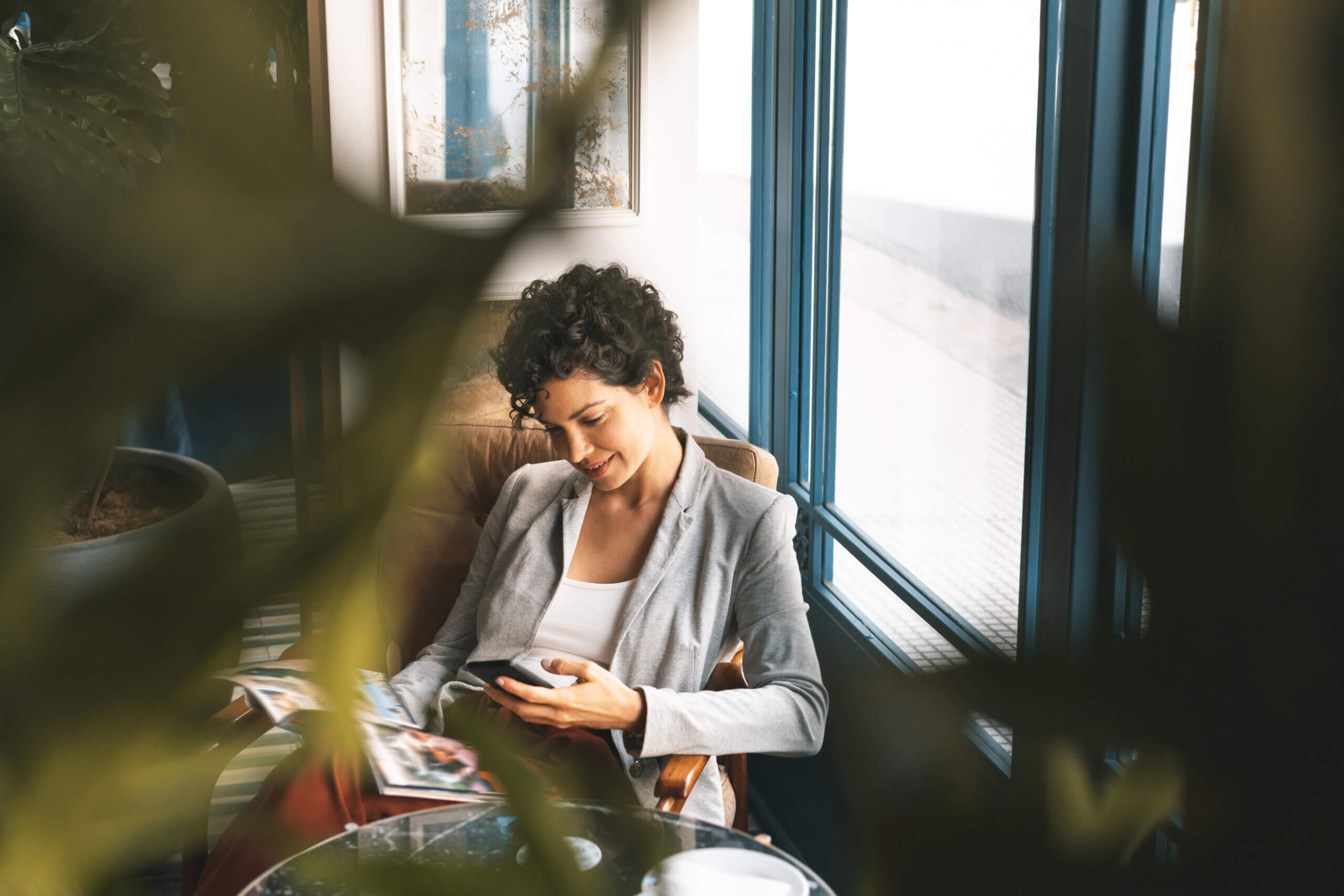 Woman looking at cellphone and sitting in a chair next to a window.