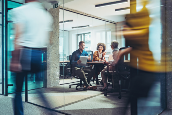 Three people sitting at a desk in a conference room with people walking around outside of it