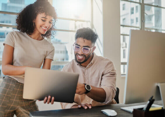 Two BIPOC young professionals - a woman standing and a man sitting - excitedly look at a tablet screen in an office setting.