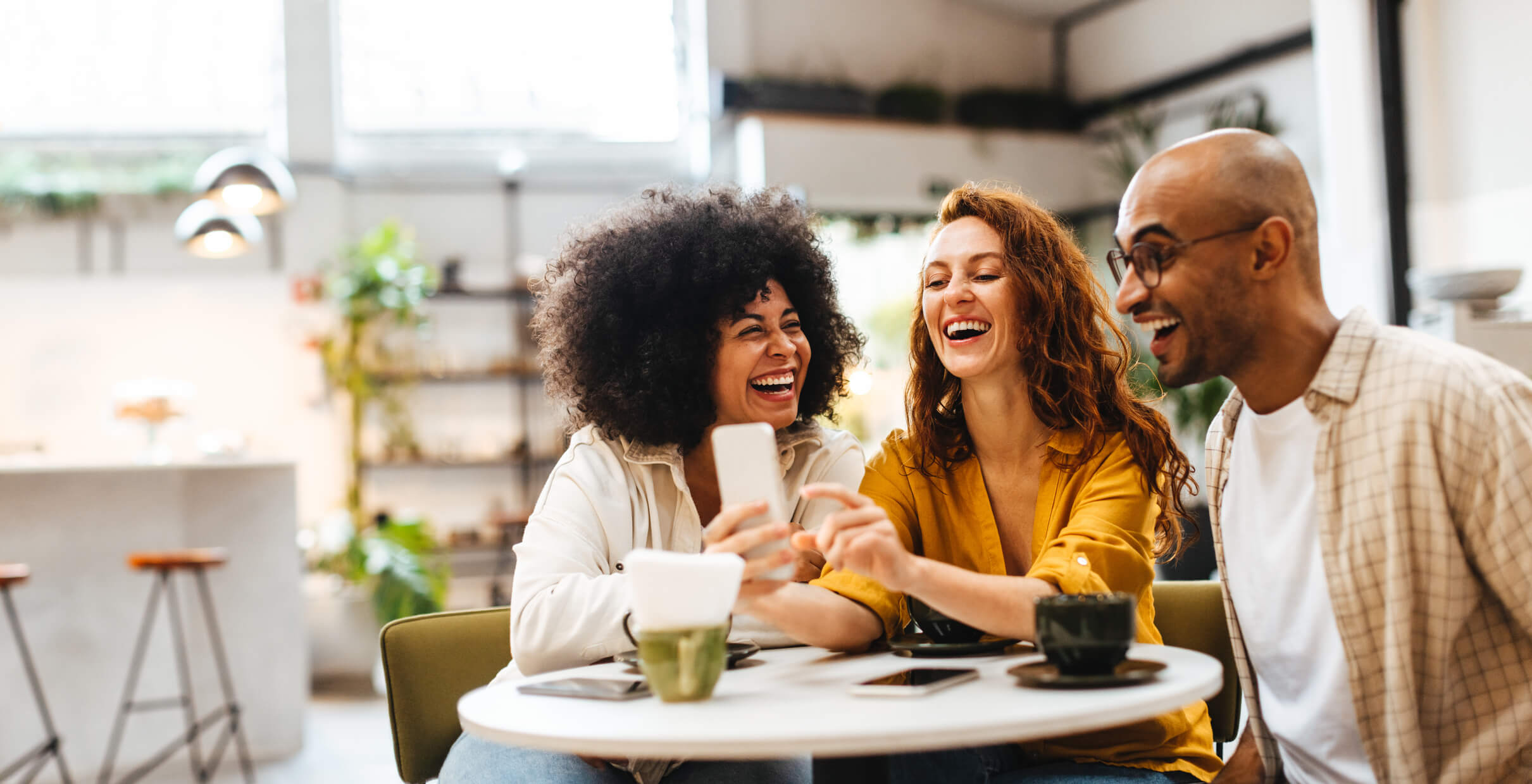 Group of happy friends browse social media on a smartphone in a lively cafe, sharing photos and updates with each other. Carefree buddies having fun as they hang out together over coffee.