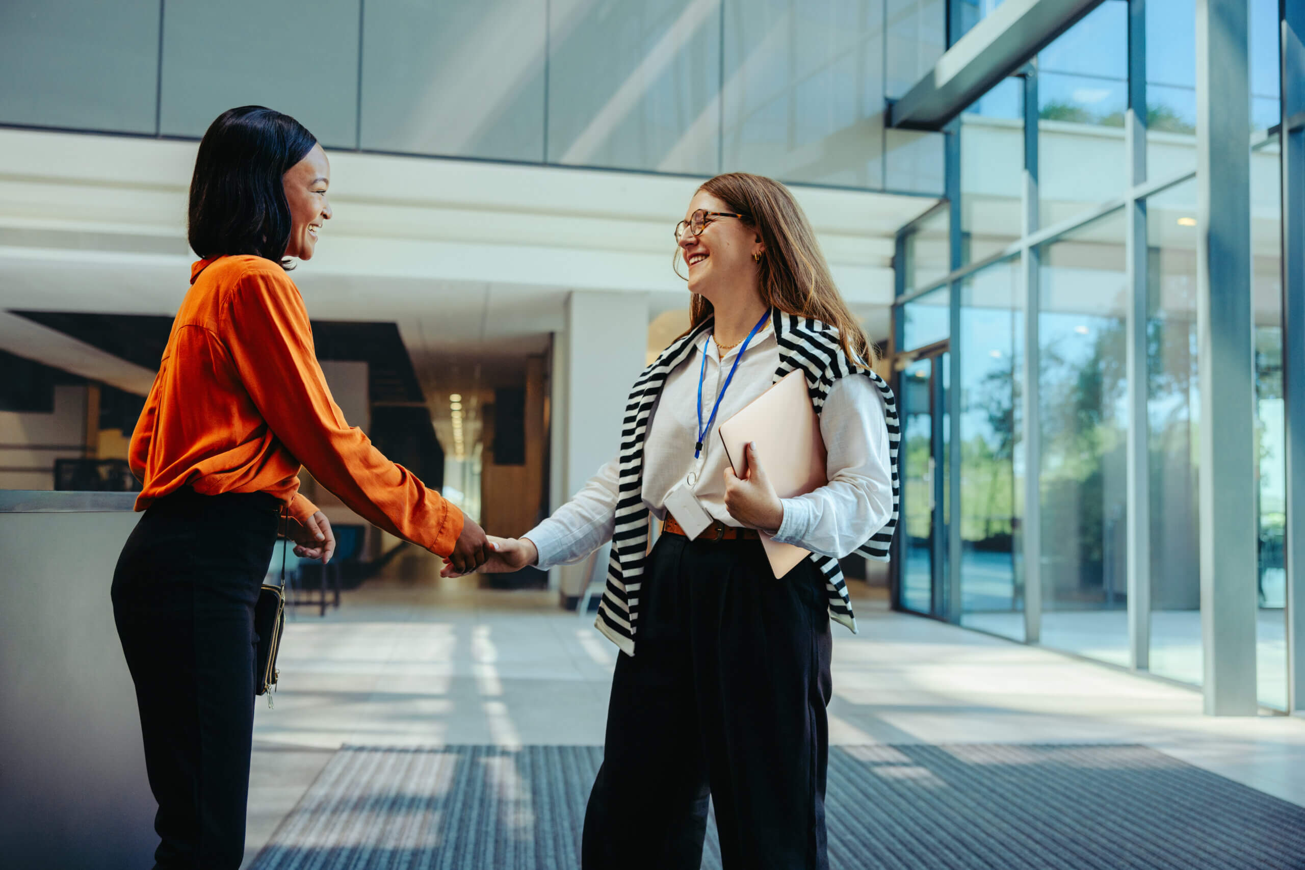 Two professional women exchanging a warm handshake at an office entrance, representing a welcoming business environment and career opportunities.