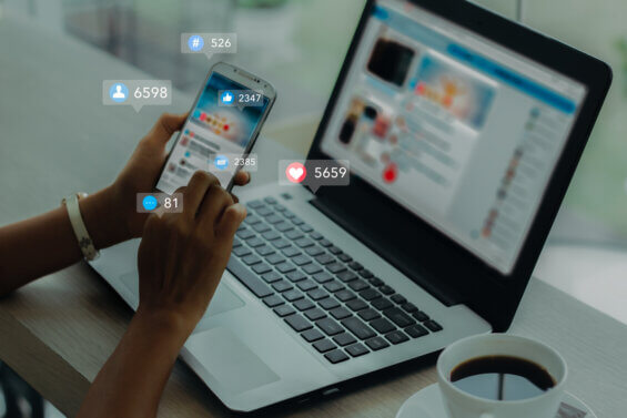 Hand holding a phone in front of an open computer with LinkedIn reaction emojis floating in the air around it.