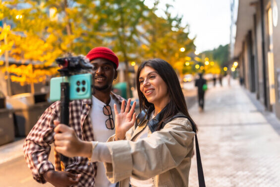 Two young adult friends creating social media content outdoors, filming a live video with a smartphone on a gimbal stabilizer while walking along a urban sidewalk